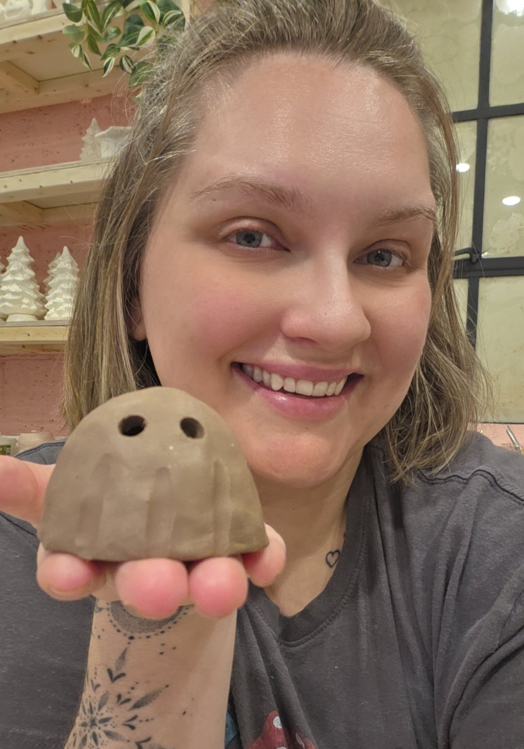 A smiling woman holding up a small, handcrafted brown clay ghost figurine with two black eyes. She is wearing a dark gray graphic t-shirt and has visible arm tattoos, set against a cozy indoor background with shelves and holiday decorations.